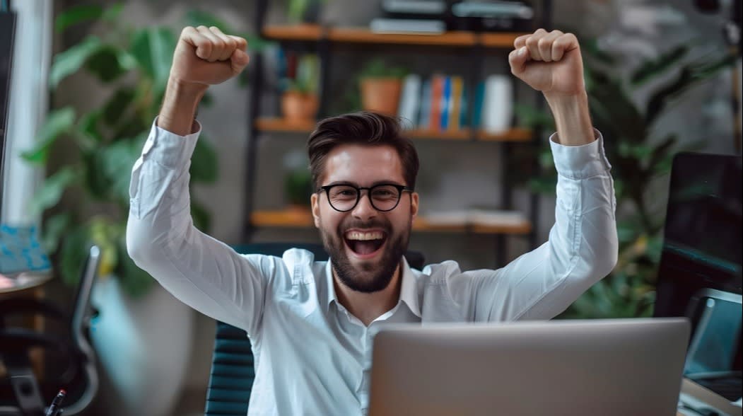 Young man celebrating success with raised arms in a home office, sitting at a desk with a laptop, bookshelves, and plants in the background.