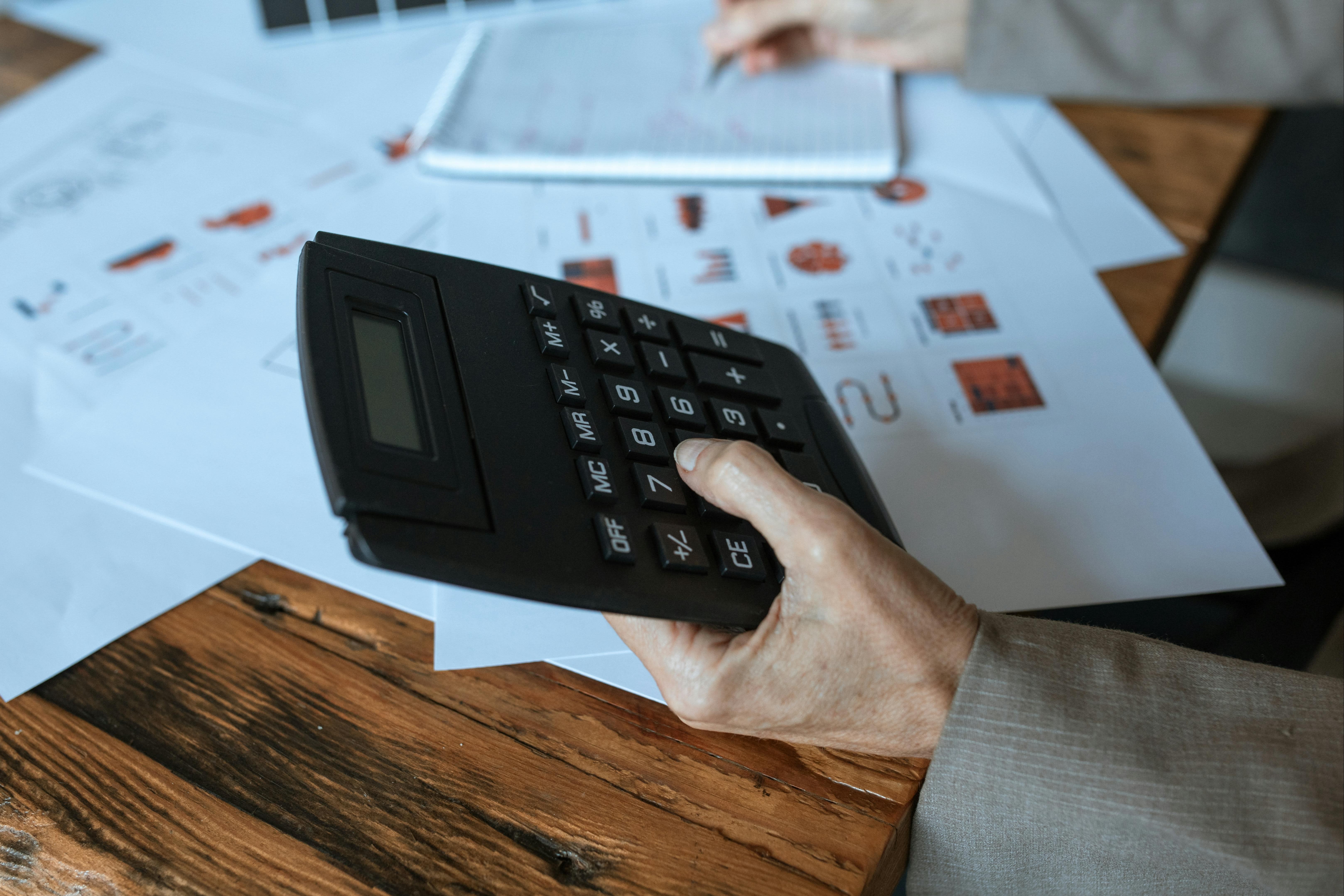 A close-up of a person's hand using a calculator with financial charts and notes spread out on a desk, indicating financial planning or analysis.
