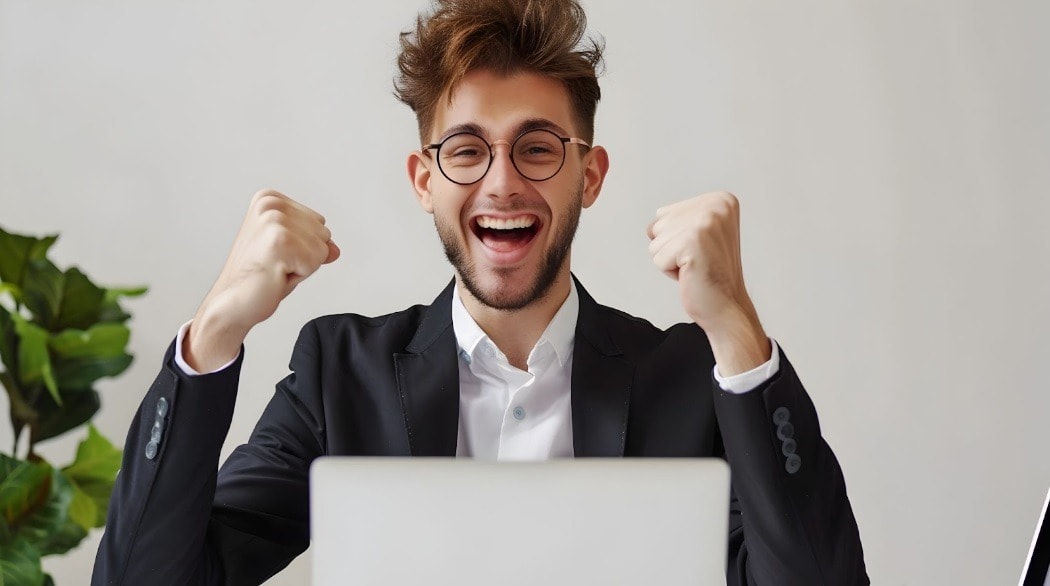 A man in glasses and a suit raising his arms in excitement while working on a laptop, symbolizing professional or academic success.