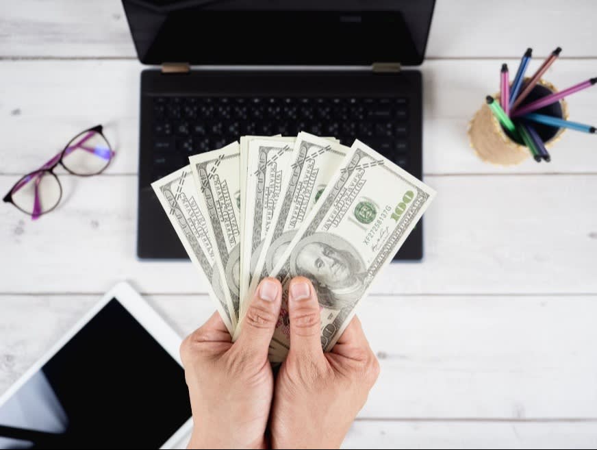 A person holding a spread of U.S. hundred-dollar bills over a desk with a laptop, tablet, and pencils, representing financial success or earnings.