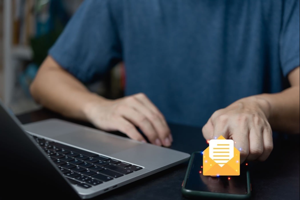 A person tapping a smartphone with a glowing email icon next to a laptop, symbolizing email communication and notifications.