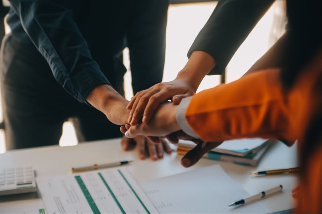 A group of hands stacked in a gesture of teamwork on a desk with documents and office supplies, symbolizing collaboration in a corporate setting.