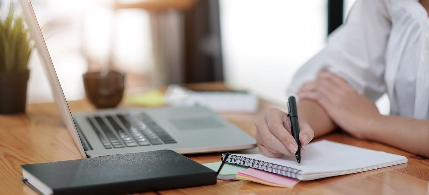 A person writing in a spiral notebook at a desk with a laptop, sticky notes, and a black notebook, in a well-lit workspace.