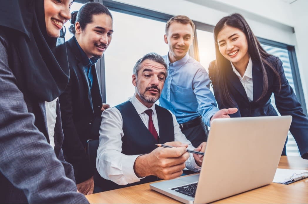 A group of business professionals in a meeting, gathered around a laptop, discussing a project or presentation, with a man leading the discussion.