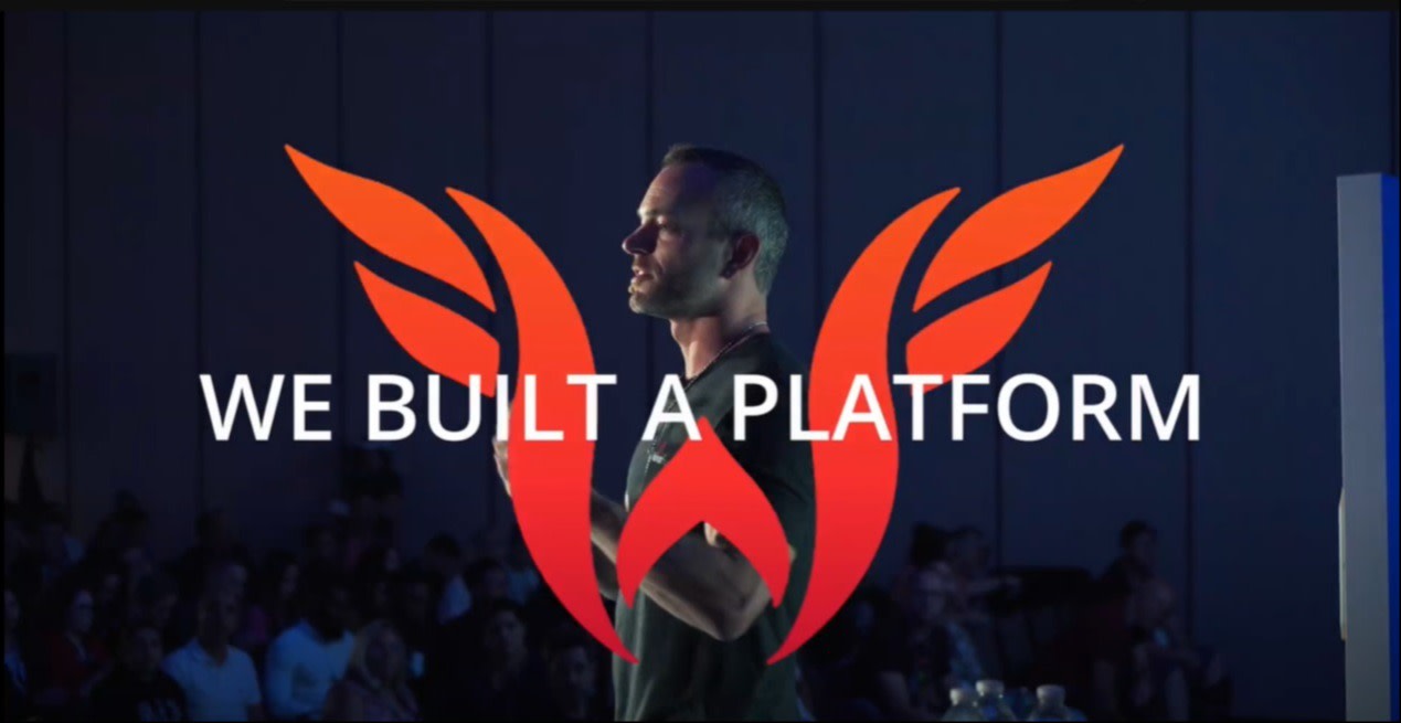 Speaker presenting on stage with the Webinar Fuel logo, featuring a bold red "W" with wings and the text "We Built A Platform" overlaid on the image.