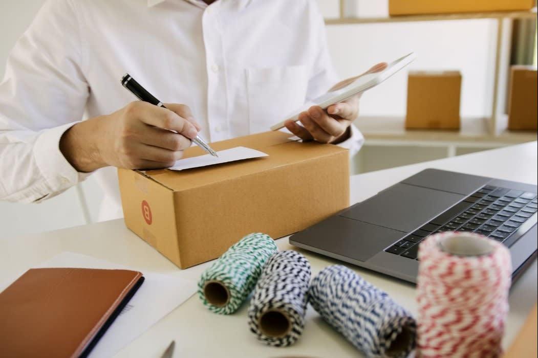 Person writing on a shipping label for a cardboard box while holding a smartphone, with a laptop and packaging supplies on a table, symbolizing e-commerce business.