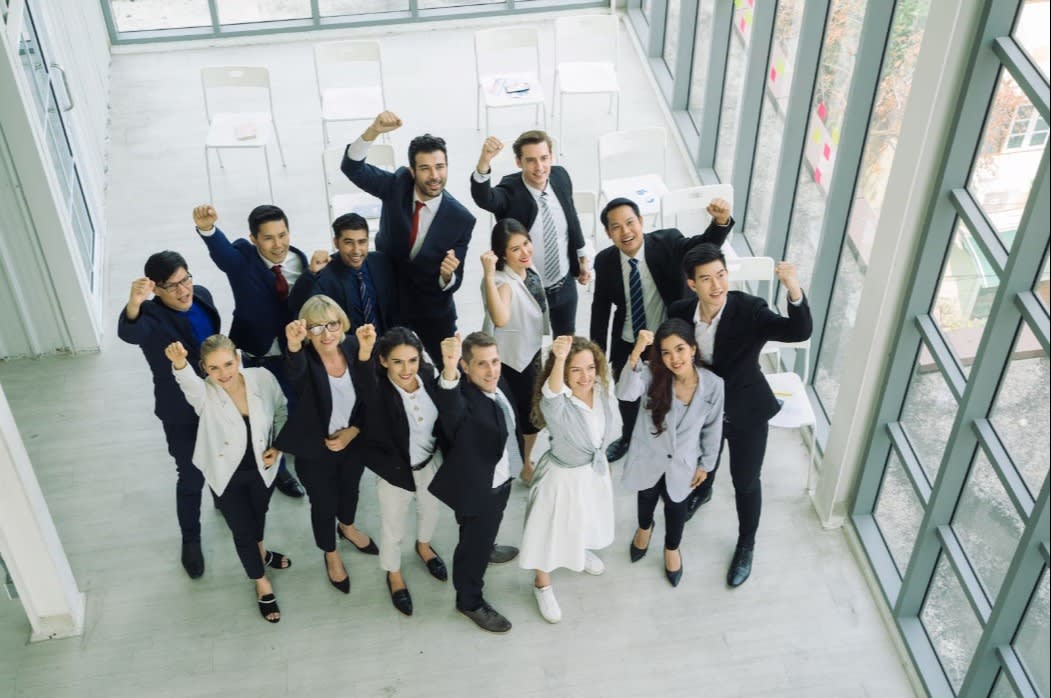A diverse group of business professionals raising their fists in celebration in a modern office, symbolizing teamwork, success, and unity.