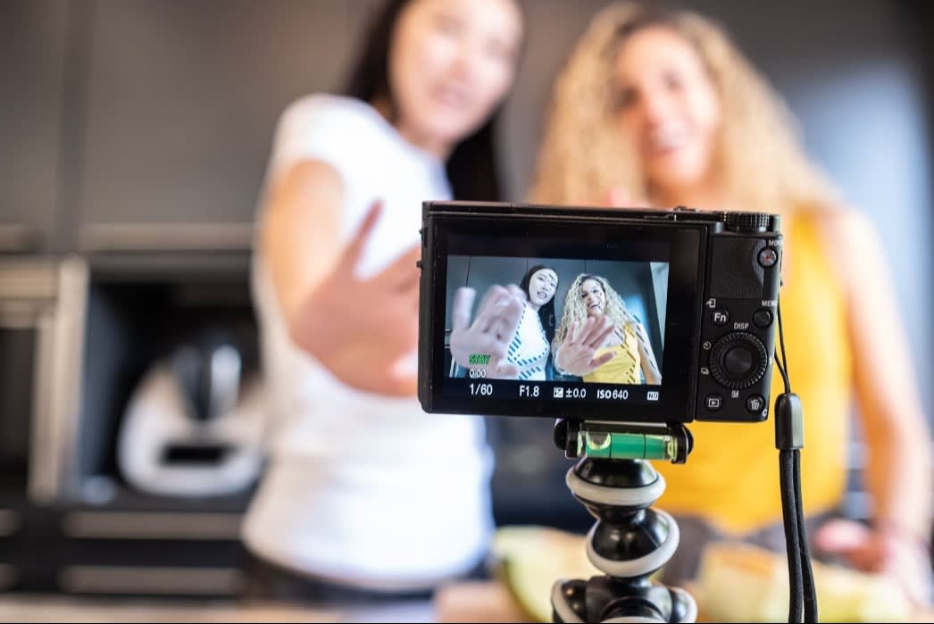 Two women recording a video with a digital camera on a tripod, shown gesturing towards the camera in the screen display.