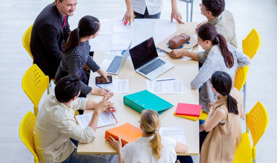 A group of colleagues collaborating around a table with laptops, documents, and colored binders, actively discussing a project in a team meeting.
