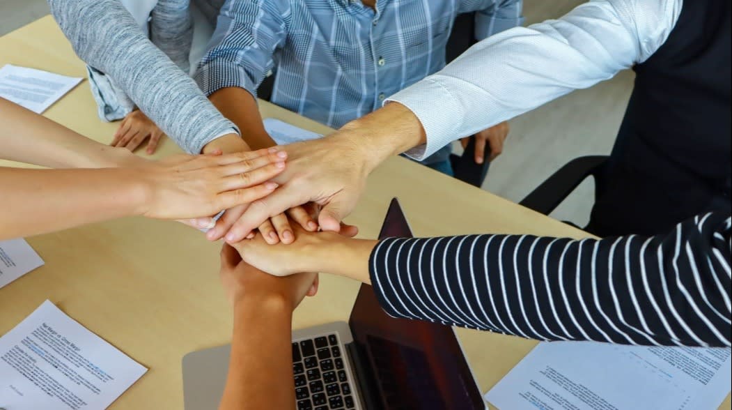 Team members placing hands together over a table with documents and a laptop, symbolizing teamwork and collaboration in a business setting.