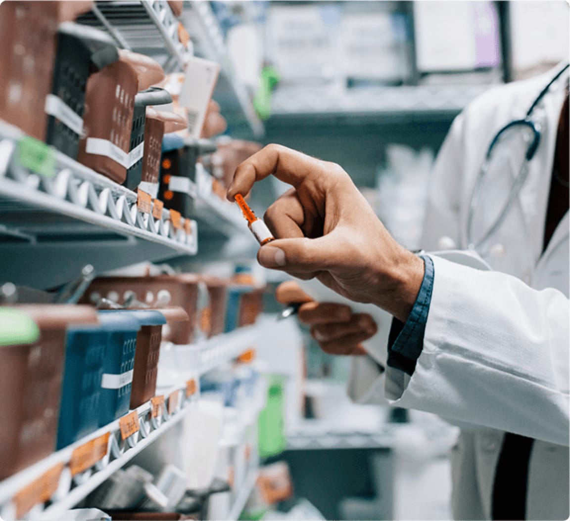 Pharmacist in white coat holding a small medication vial while selecting from organized pharmacy shelves