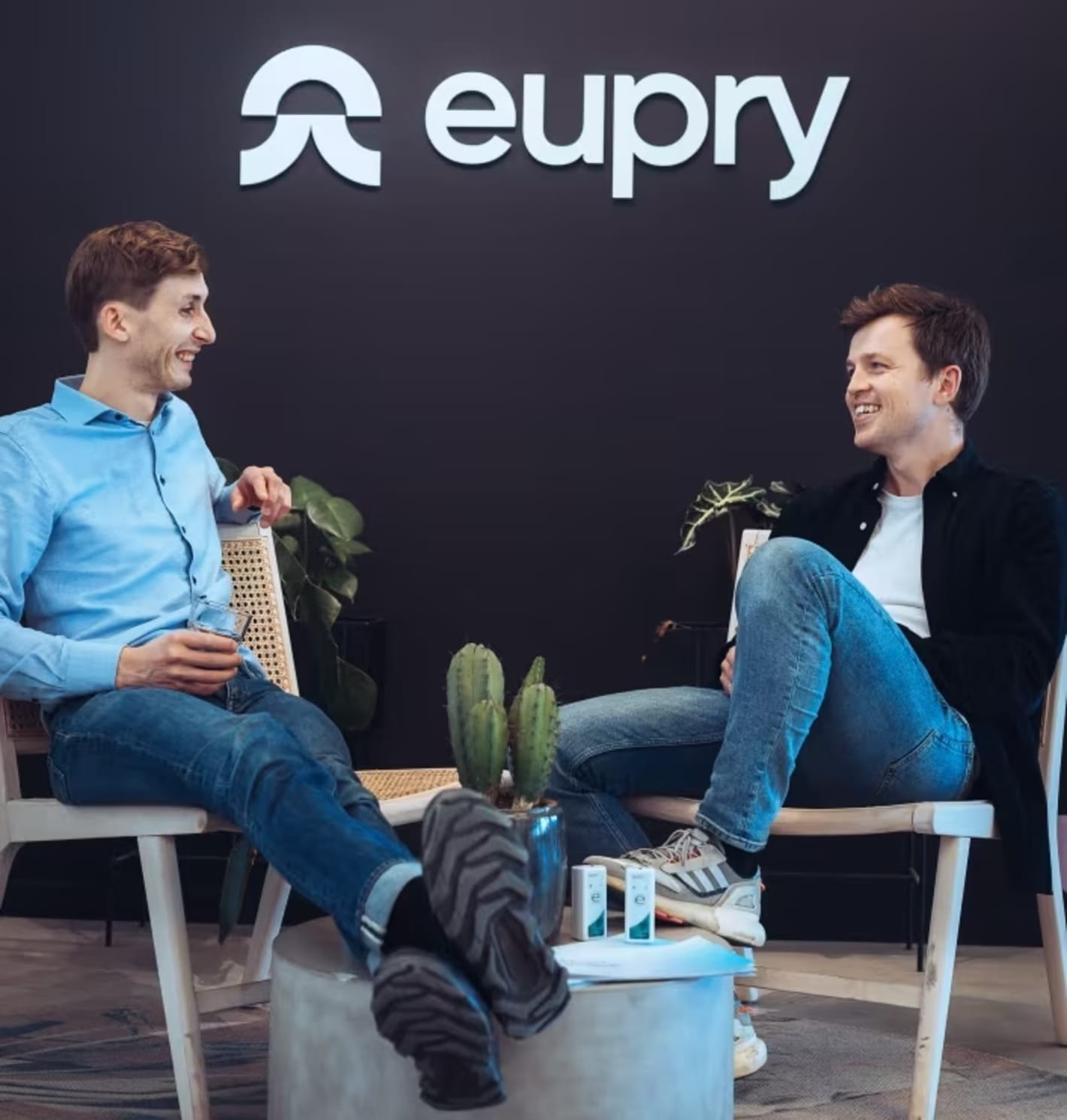 Two men sitting in chairs laughing during a conversation in front of the Eupry logo on a dark wall.