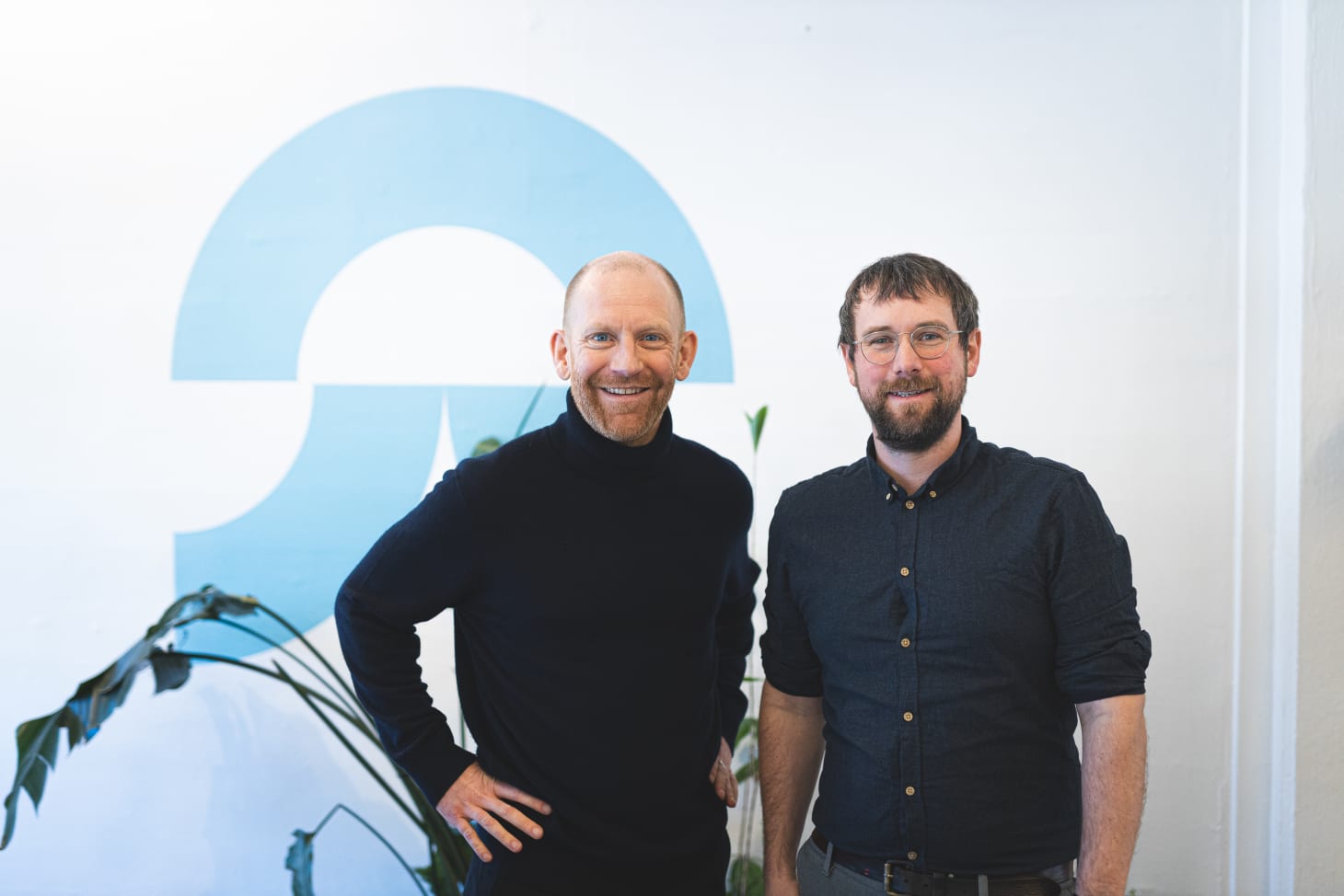 Two men smiling in front of a blue Eupry logo on a white wall, with indoor plants nearby.