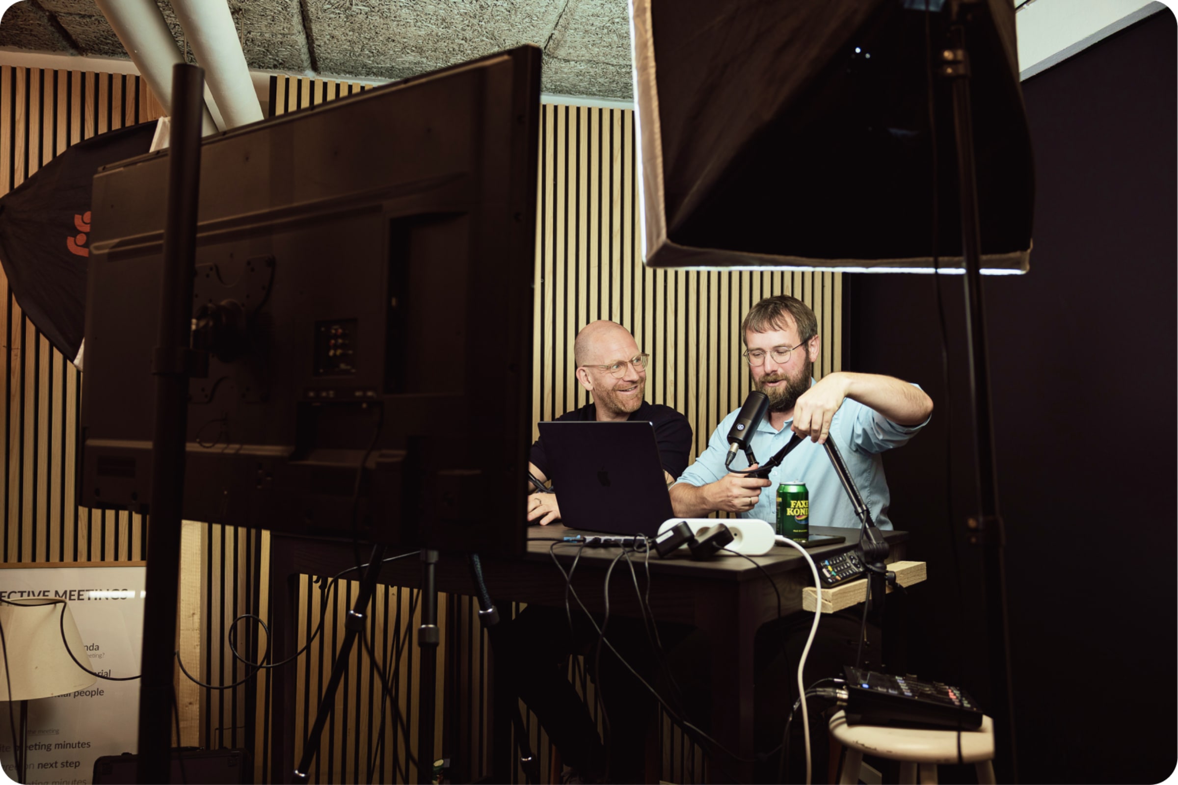 Two men recording a podcast in a studio setup with microphones, laptop, lighting equipment, and a green beer can on the desk.