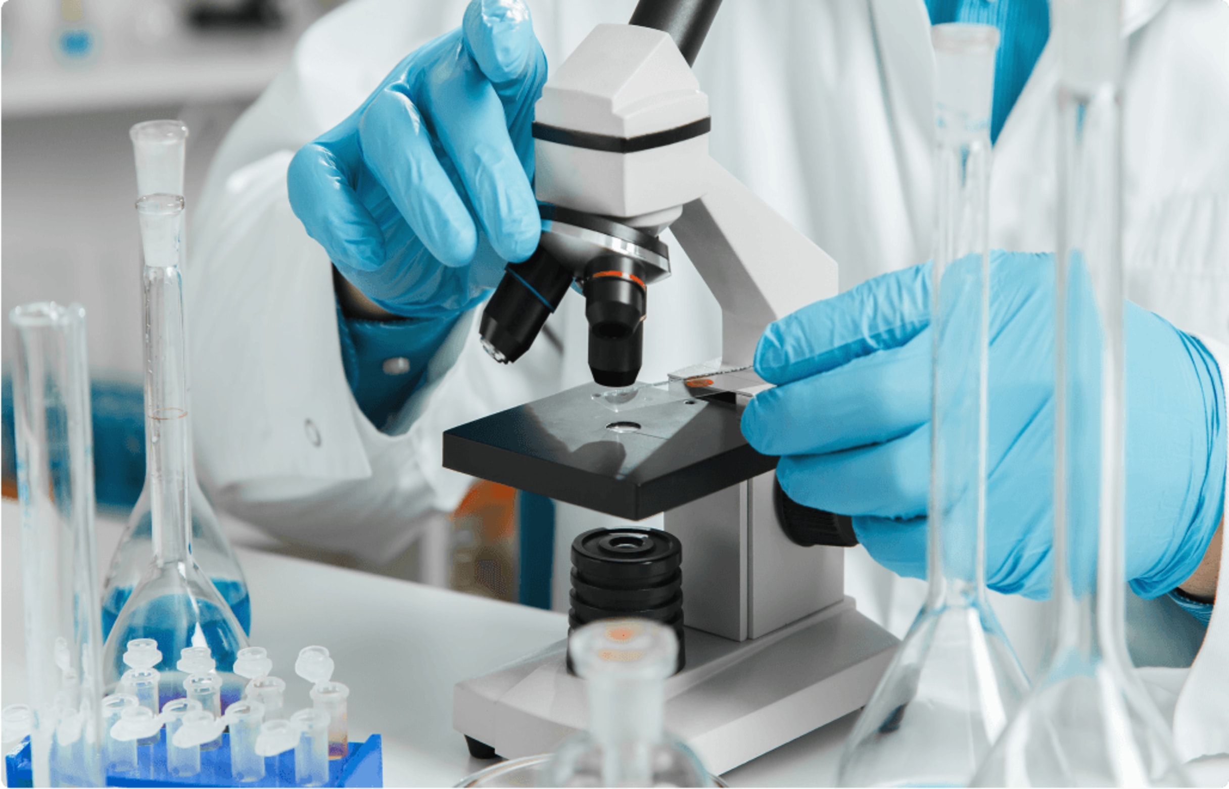 Scientist in white lab coat and blue gloves placing a slide under a microscope surrounded by laboratory glassware