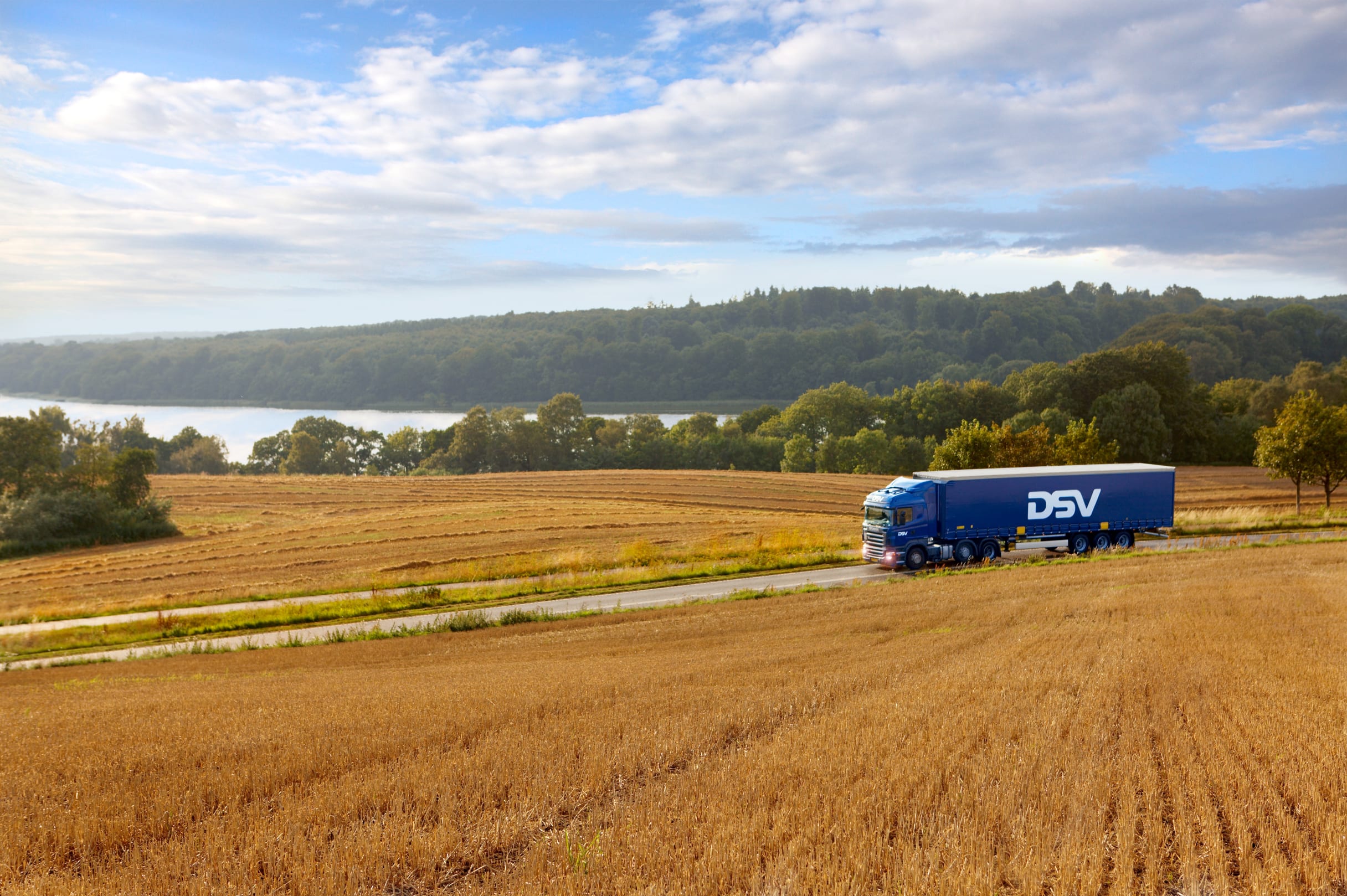 DSV blue semi-truck driving through golden harvested fields with a lake and forest in the background