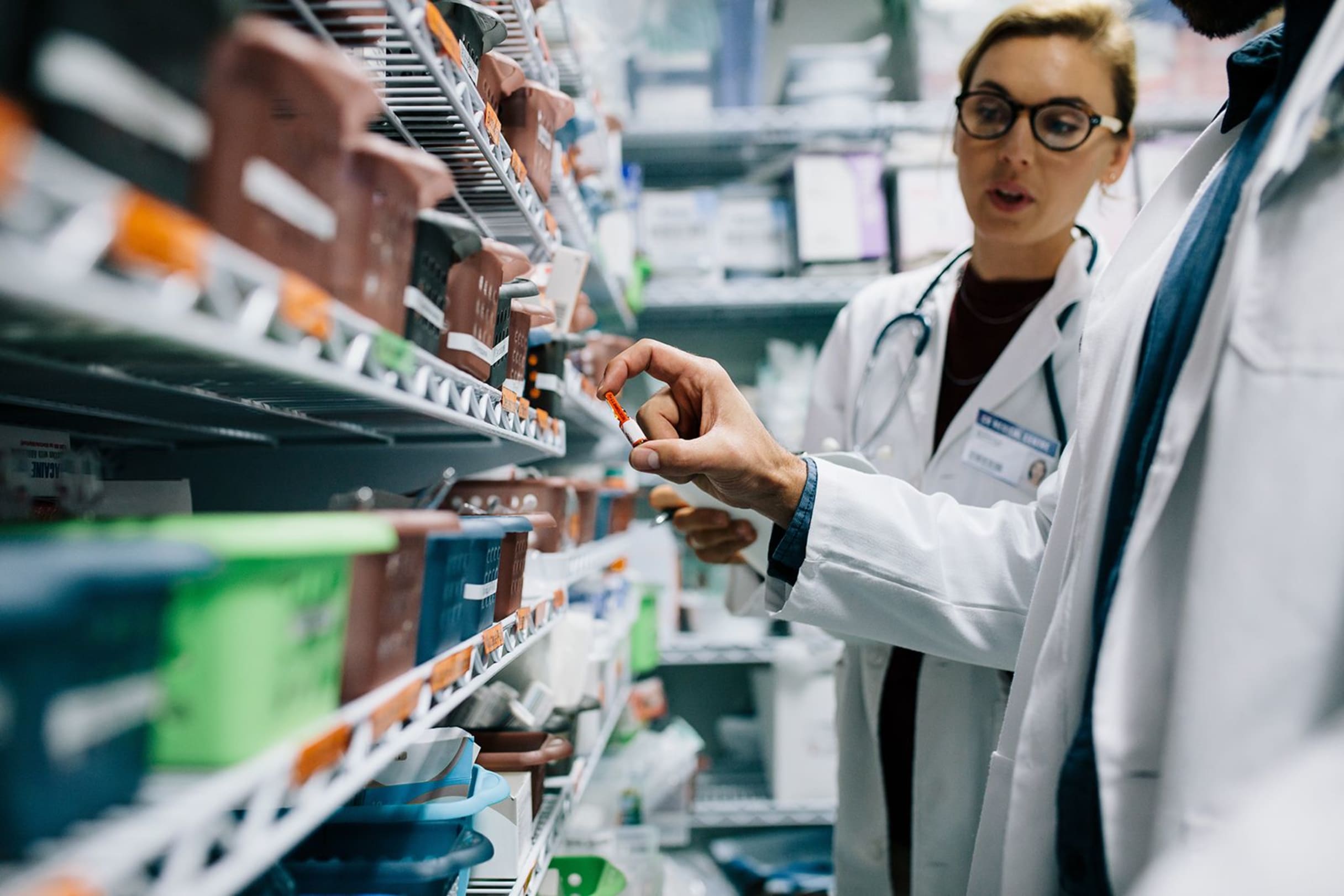 Pharmacist in white coat and stethoscope examining medication vial on stocked pharmacy shelves