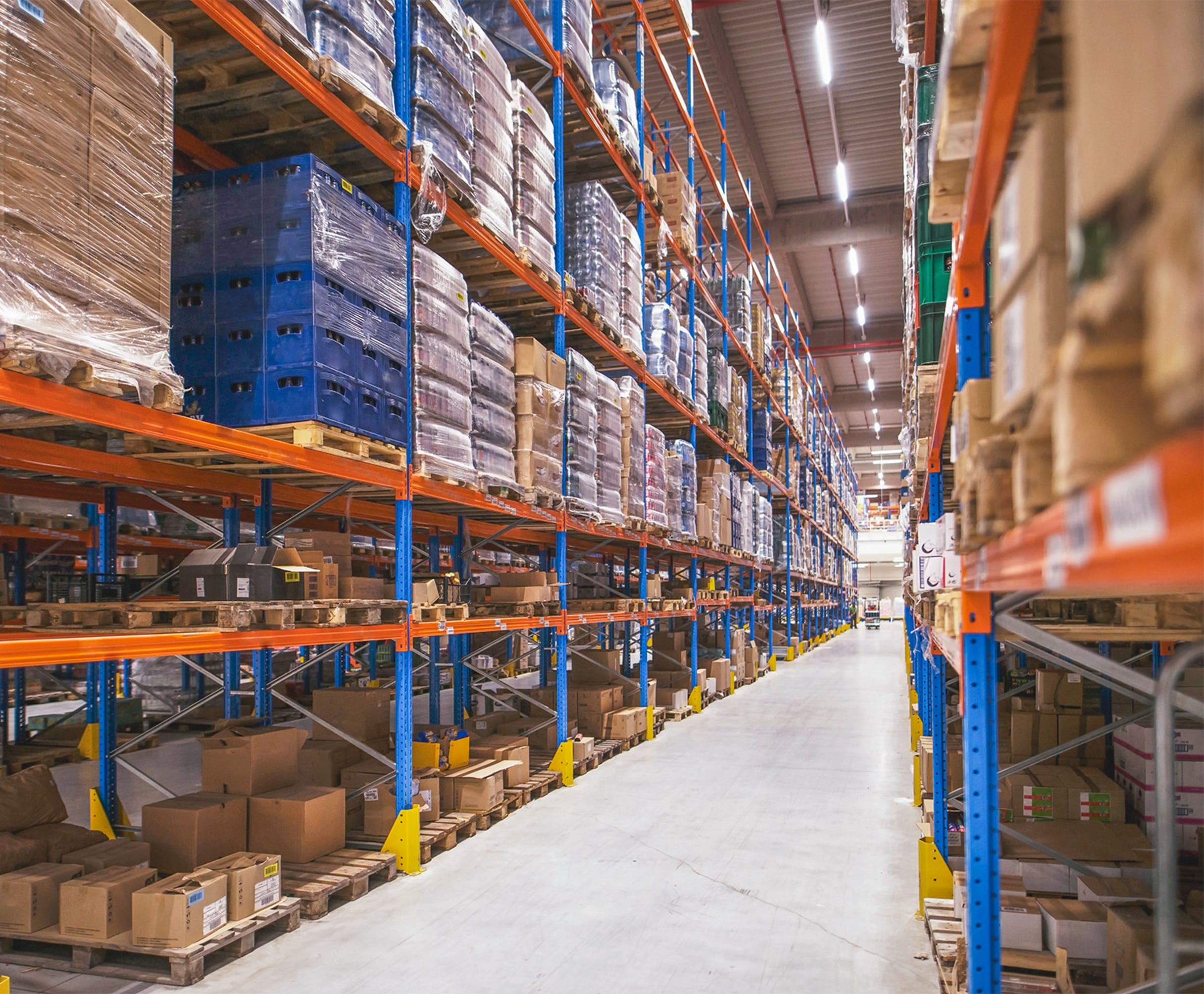 Large-scale warehouse aisle with tall blue and orange shelving racks stocked with palletized goods and cardboard boxes.