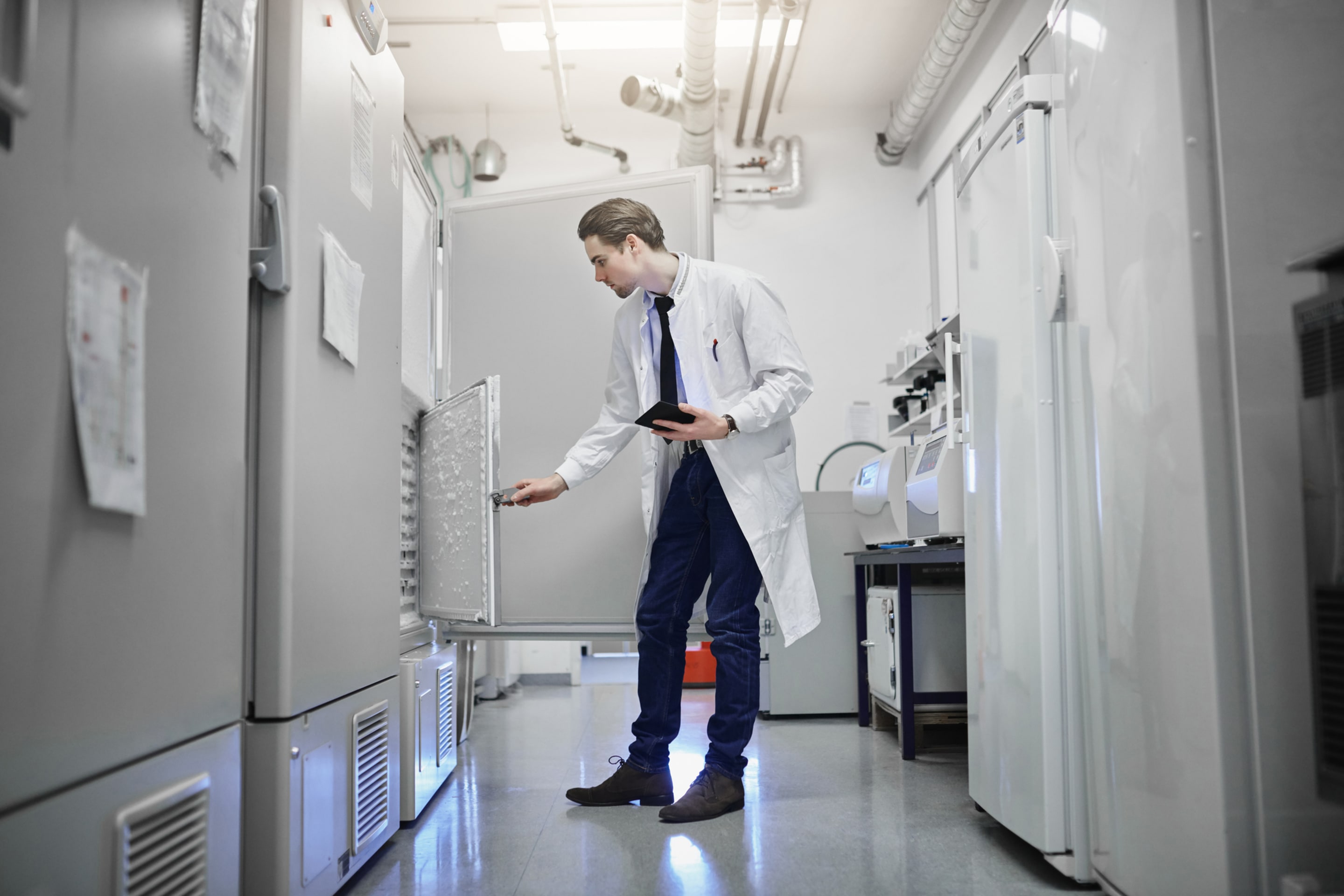 Lab technician in white coat checking ultra-low freezer with tablet in pharmaceutical storage room
