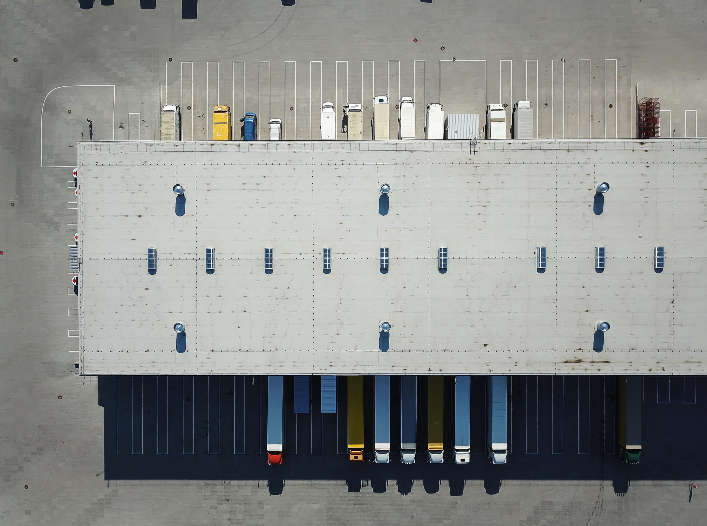 Aerial view of a logistics warehouse with trucks docked at loading bays and parked vehicles on the rooftop lot.