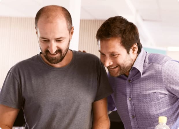 Two men smiling and leaning in together, looking at something off-camera in a bright office setting.
