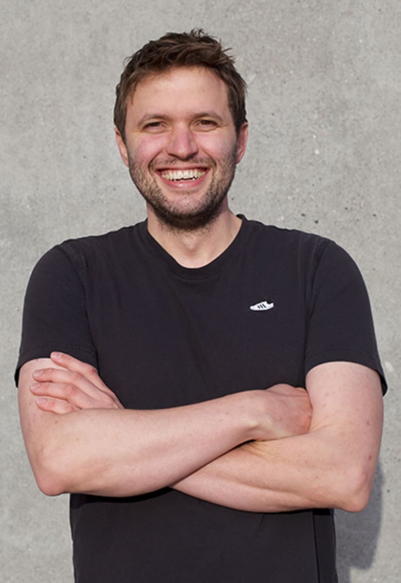 Smiling man with crossed arms wearing a black Adidas t-shirt, standing against a gray concrete wall.