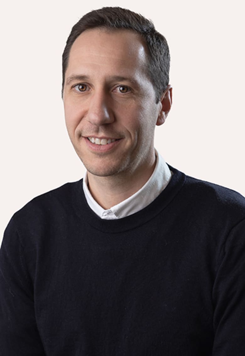Headshot of Christian Holse smiling, wearing a dark navy sweater over a white collared shirt, light background.