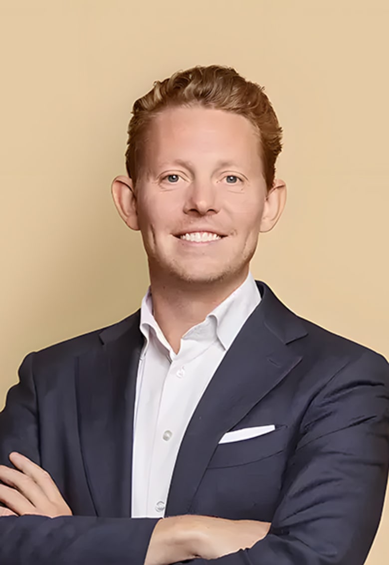 Professional headshot of Ditlev, smiling man in navy blazer with auburn hair, arms crossed, beige background.