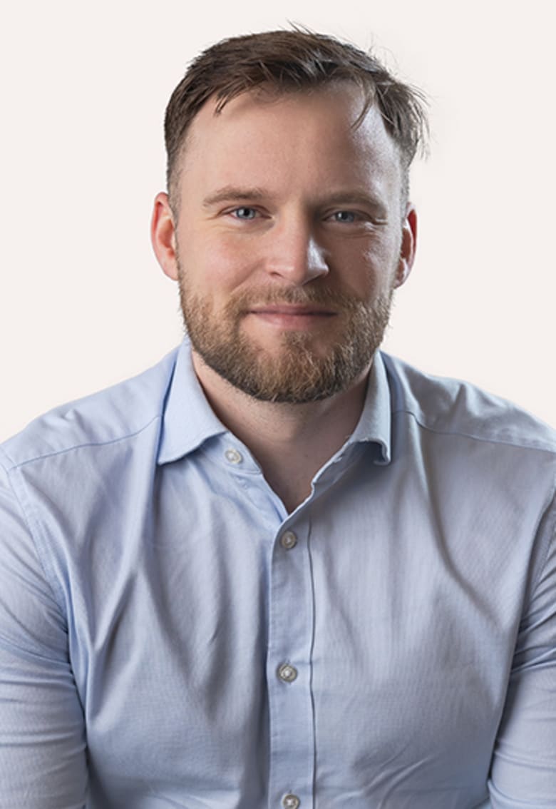 Smiling man with short brown hair and beard wearing a light blue button-up shirt against a white background