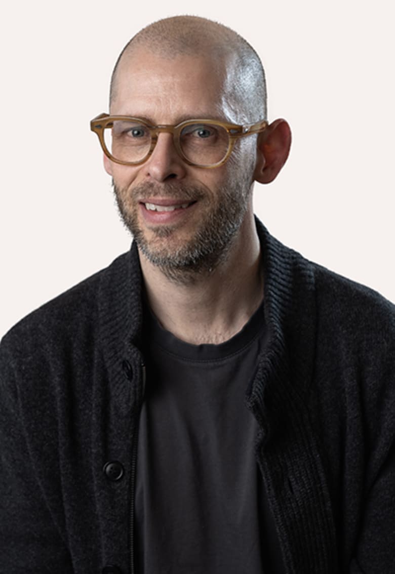 Bald man with amber glasses and short beard, wearing black cardigan over dark t-shirt, smiling at camera