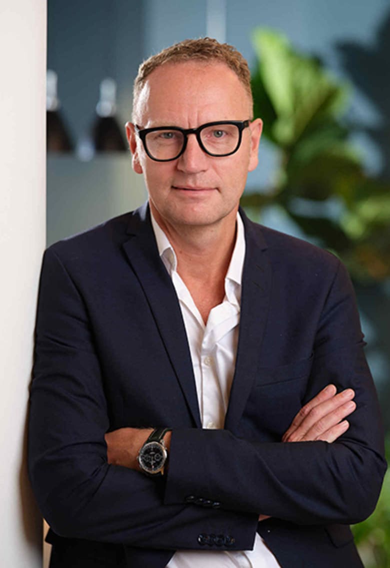 Middle-aged man in navy blazer and glasses, arms crossed, posing professionally indoors with green plants in background.