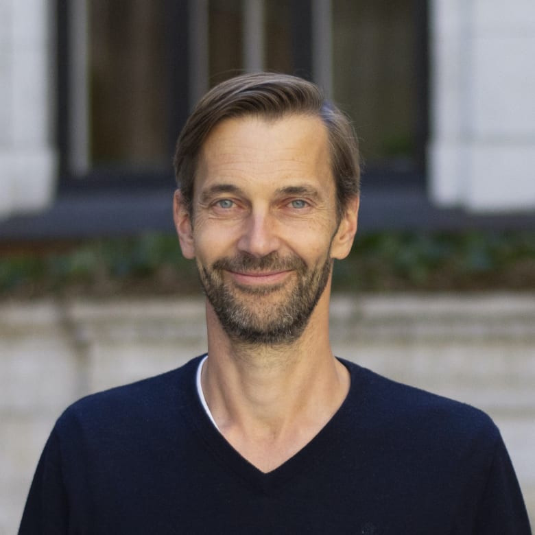 Portrait of a smiling man with beard in navy V-neck sweater, standing outdoors near windows.