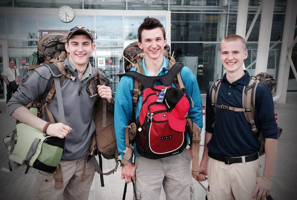 Three young men with backpacks smile for the camera at an airport.