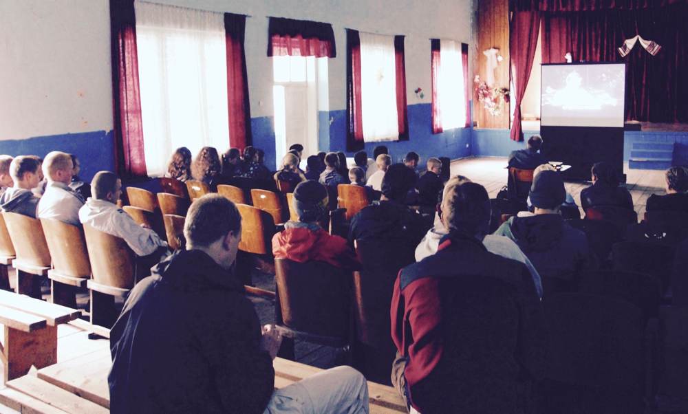 A village audience sits in rows of wooden seats, watching a film projected at the front of a hall.