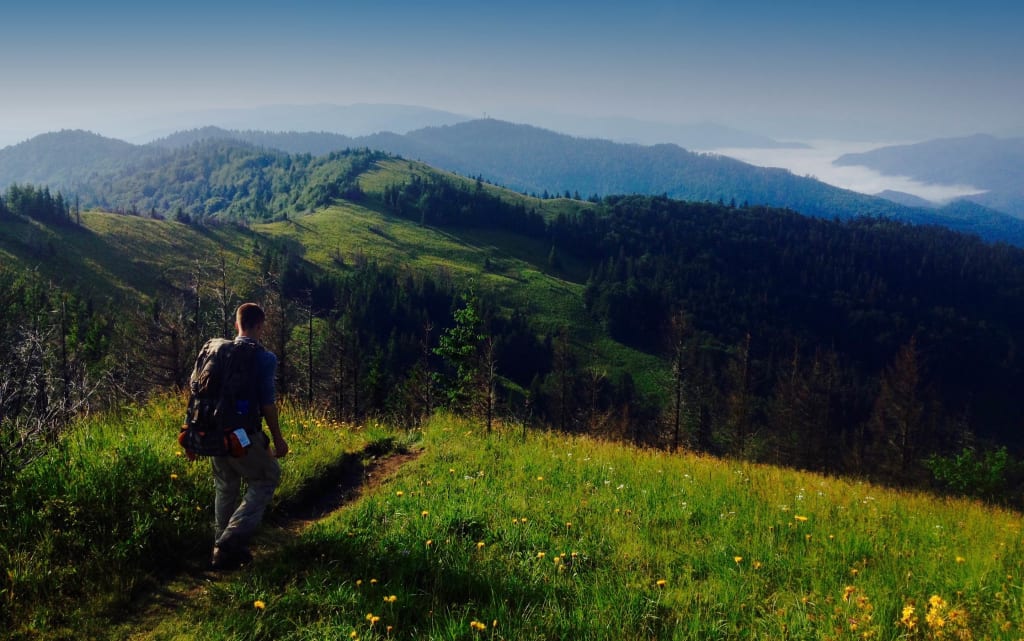 A hiker with a backpack walks a grassy ridge above forested mountains and valleys of mist.