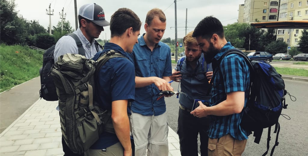 A team of young men with backpacks gathers around a map on a street in Ukraine, planning the day's route.
