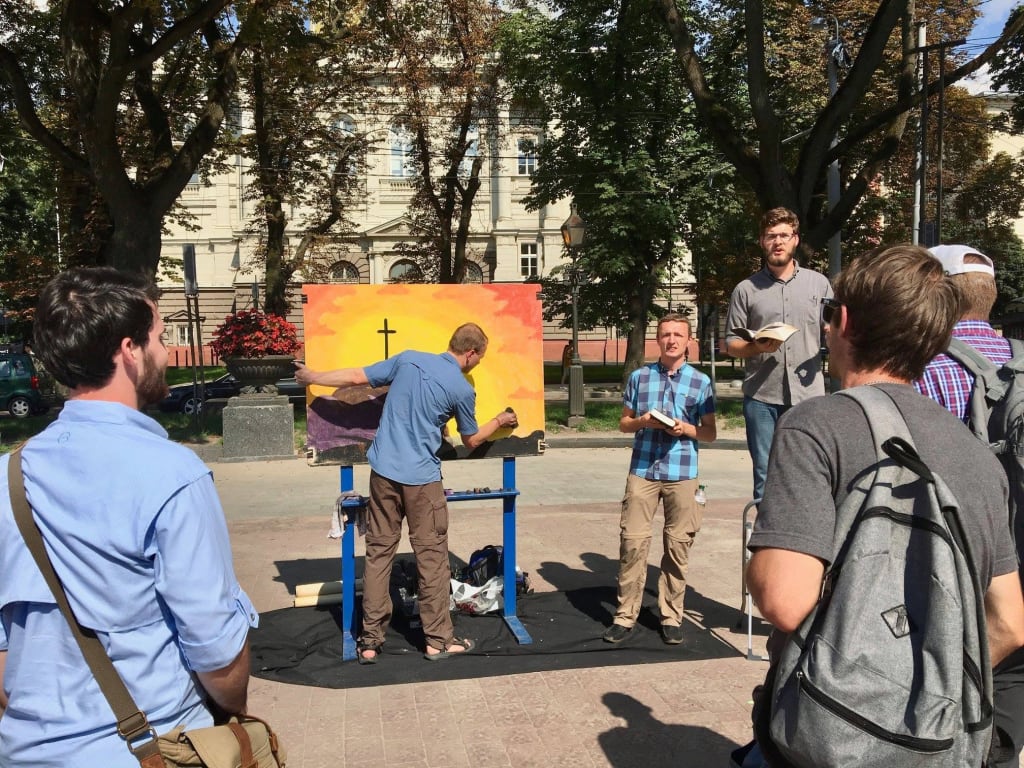 A man paints a cross against a sunset on a large easel in a city square while another preaches from an open Bible and a small crowd listens.