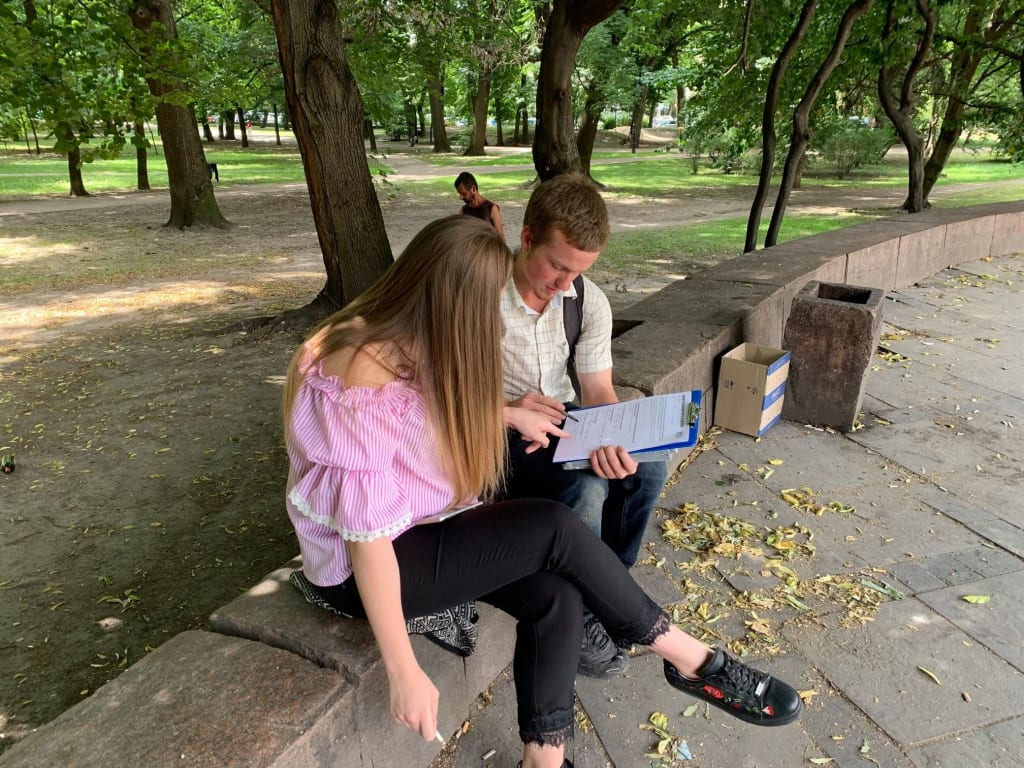 A young man and a young woman sit side by side on a low park wall, reading through a survey on a clipboard.