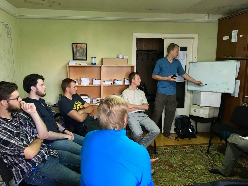 A group of young men sit around a room listening as a man explains something on a whiteboard.