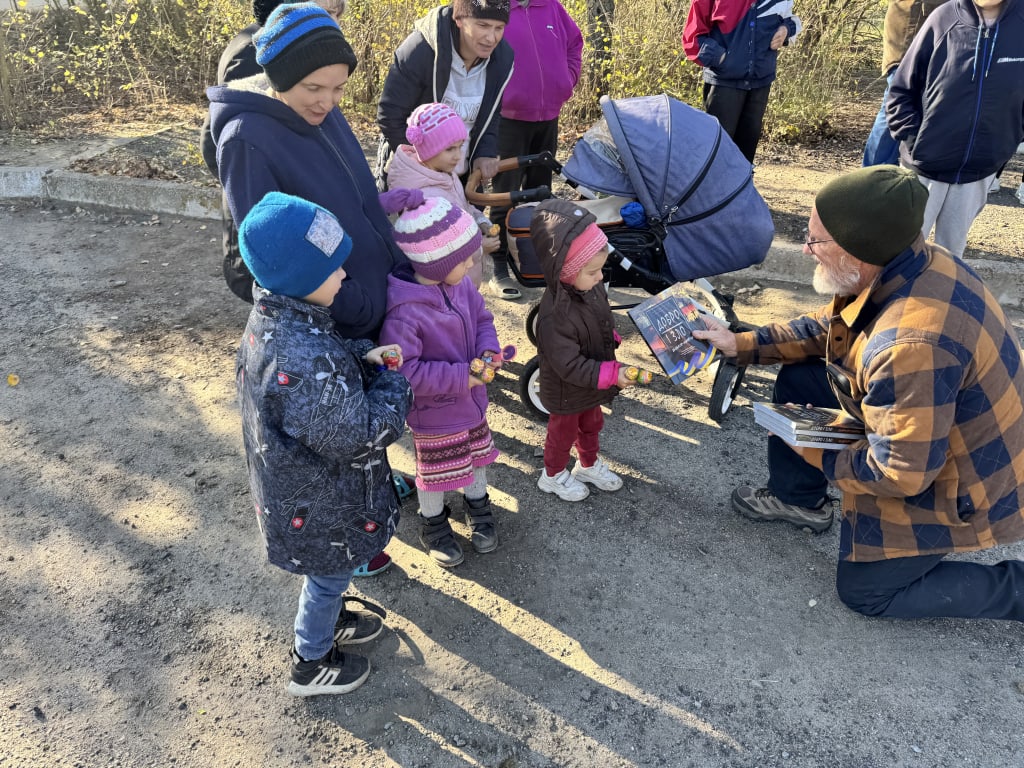 Ben Sargent crouches to hand a book to a small child in a winter coat.