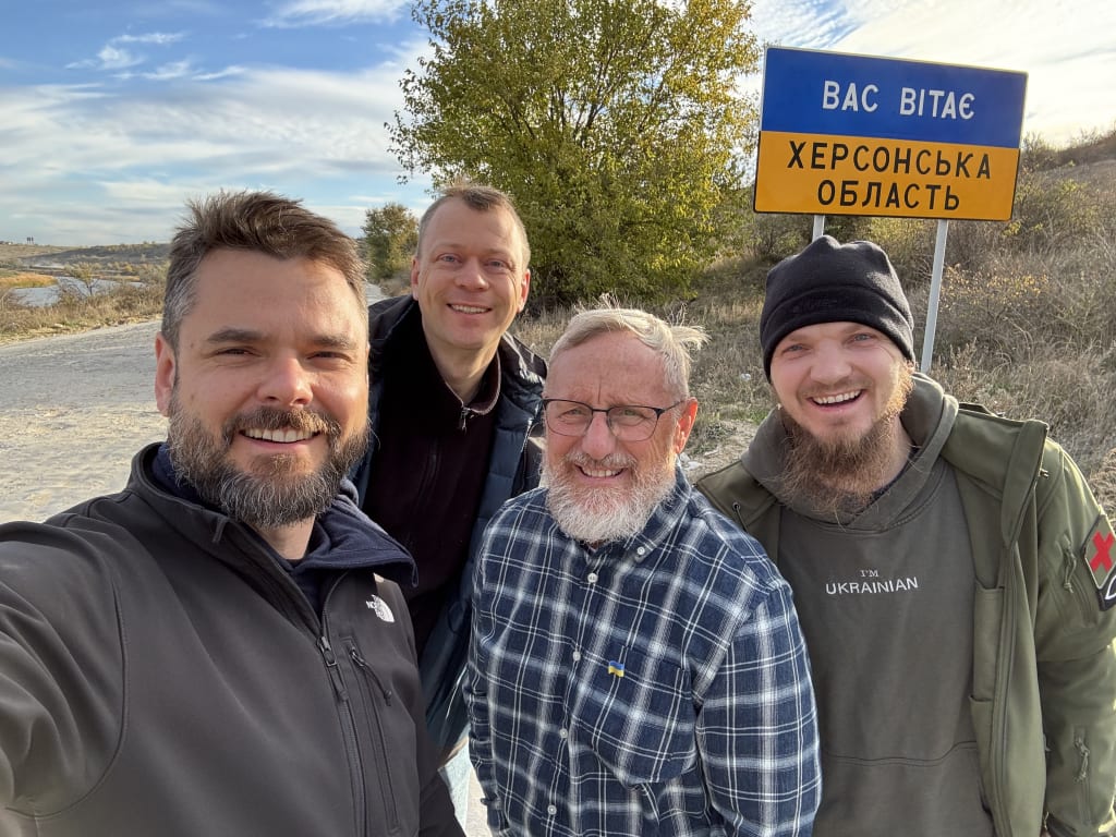 The team and a Ukrainian partner pose in front of the blue-and-yellow Kherson Oblast border sign.