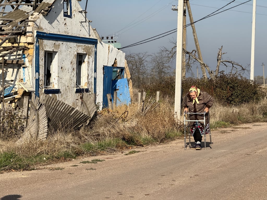 An elderly woman walks with a walker past a ruined house.