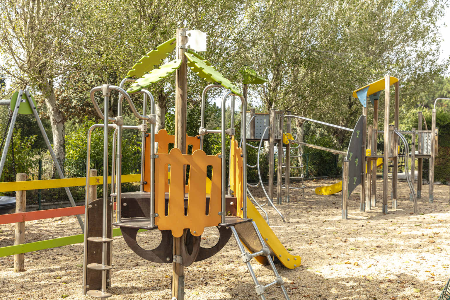 Metal and wood framed climbing frame with slide, for younger children, in the play area with sandy surface, backed by trees, at le Mane Guernehue campsite Brittany