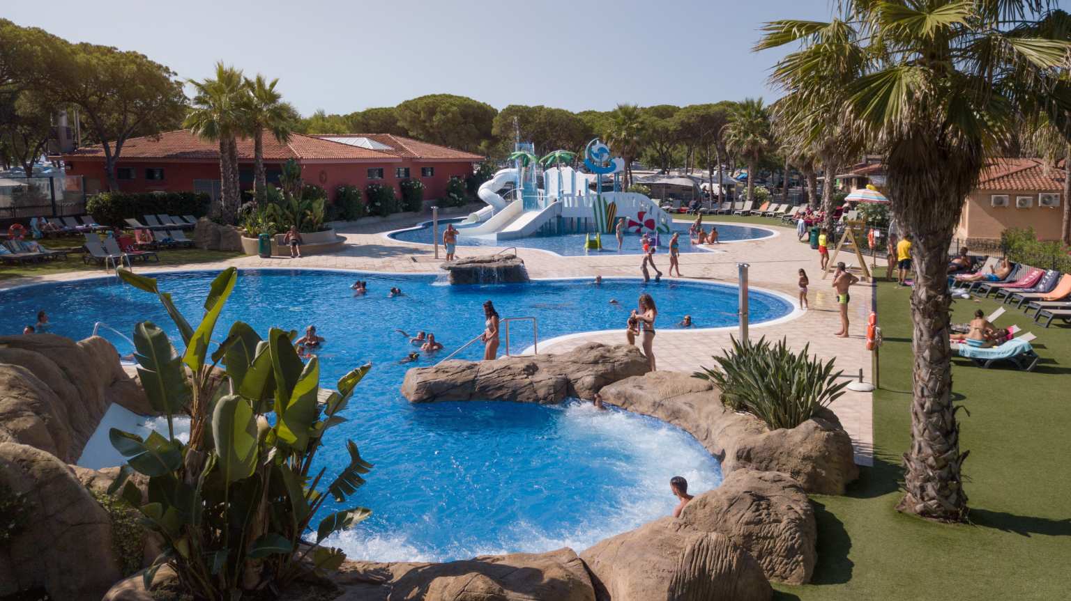 View of the lagoon pool and sunbathing lawn to the left with palm trees, looking down to kids splash pool with slides, at Bella Terra campsite, Costa Brava