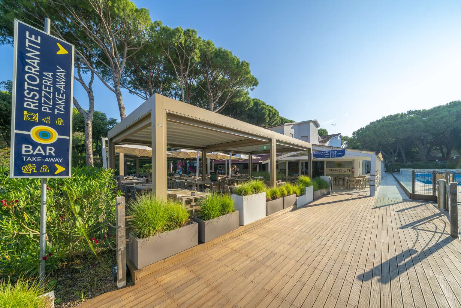 Sign for restaurant, pizza and take away, next to planters, awning and tables at Jesolo Mare Camping Village, Adriatic