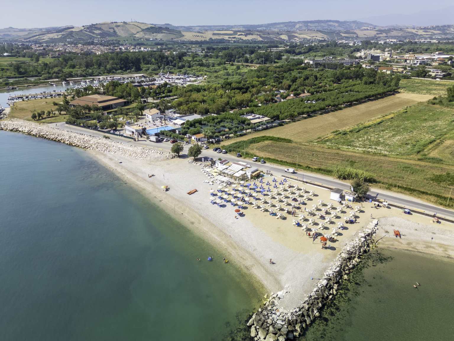 Aerial view of sunloungers and parasols on the beach next to Roseto degli Abruzzi campsite, fields, trees,houses and hills in the distance, Abruzzo, Italy 
