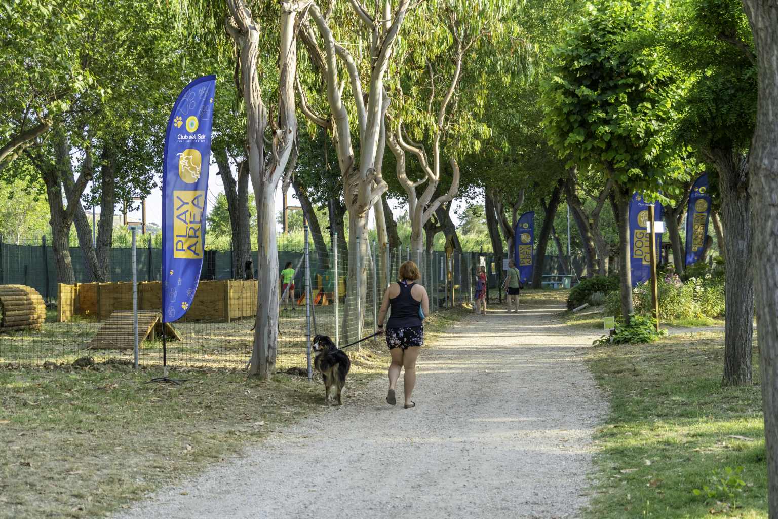 Owner walking dog next to sign to fenced dog play area with wooden activity course under trees, at Roseto degli Abruzzi campsite, Abruzzo, Italy