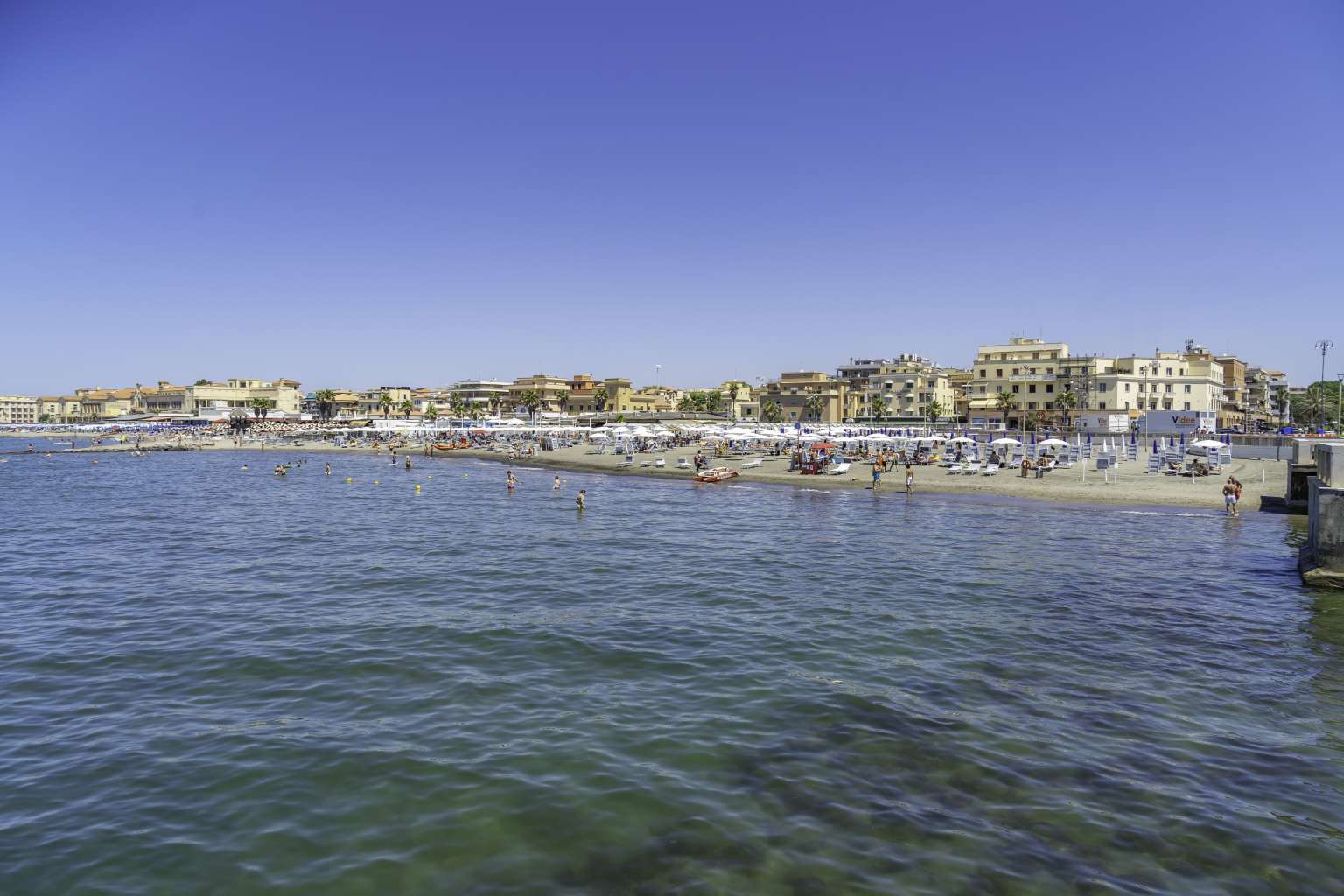 View from the sea onto Lido di Ostia beach - narrow strip of sand filled with people and sunloungers, backed with low houses, near Village Roma Capitol campsite, Rome