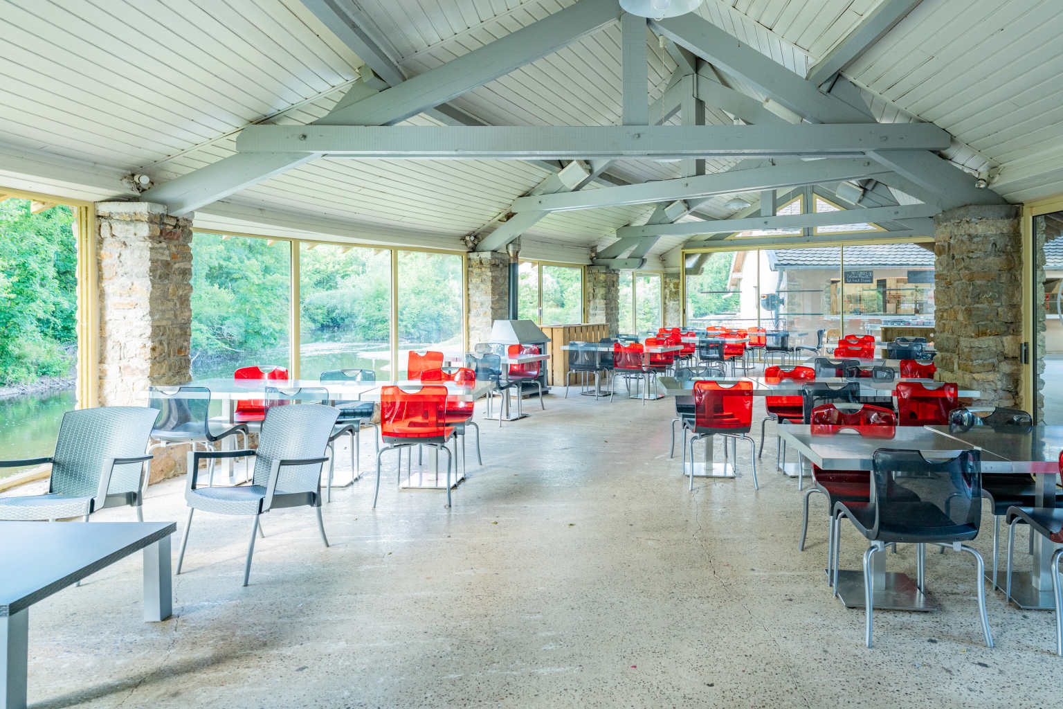 Tables and chairs next to wall to wall windows under high beamed ceiling in the onsite restaurant at Val de Bonnal campsite in the Jura