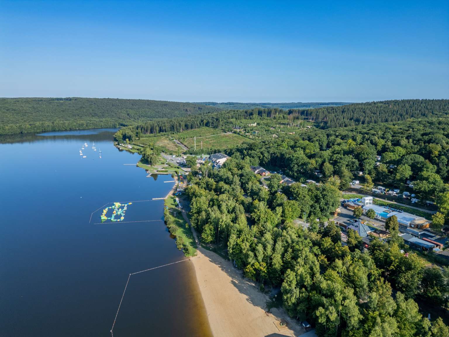 Aerial view of the pool complex set within the forrest, the lake beach, and lake inflatables at Lac des Vielles Forges campsite in the Ardennes